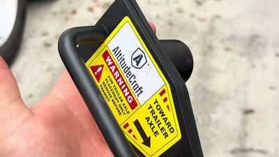 Hand holding the AltitudeCraft tandem slide stopper with its yellow warning label, toward trailer axle arrow visible